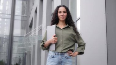 Lady businesswoman 30s director leader boss manager freelancer realtor female worker girl with curly long hair woman seller standing in city looking at camera holding laptop e-commerce online sales