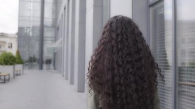 Closeup back view from behind lady businesswoman female girl with curly long hair brunette woman walking going moving in city near downtown company turns happy smiling toothy turning smile cheerful