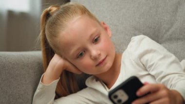 Relaxed blonde child little girl blogger cute kid teen resting on couch playing online games on mobile phone posting photos in social media apps chatting with friends watching funny cartoons at home