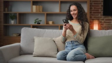 Smiling Hispanic Caucasian 30s woman housewife girl sitting on couch cozy sofa at home using smartphone for ordering food delivery chatting with mobile app looking at phone screen with smile browsing
