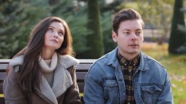 Cheerful caucasian couple lovers family sit closely on bench in autumn urban park young handsome guy yawn and hug beautiful pretty girl feel romantic embarrass on first dating rest together outdoors