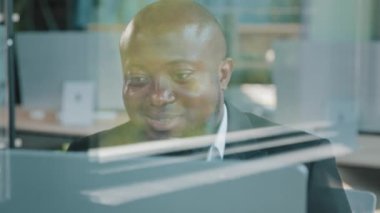 Thick bearded african male business man investor ceo analyst sit behind glassy transparent wall in office typing on computer develop internet project trade commercial data search online information
