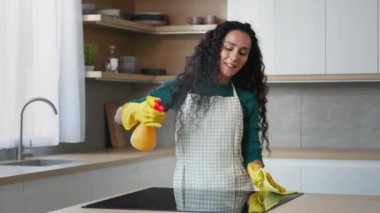 Happy young housewife arabian woman domestic cleaner maid in yellow gloves cleaning apartment washing oven by rag splashing detergent spray dancing rhythmic moves to favorite music enjoy house chores