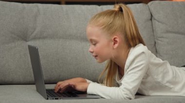 Carefree little kid pretty blonde girl child daughter using laptop lying on grey sofa surfing internet chatting with classmates playing games studying online at home. Children tech addiction concept