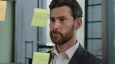 Creative bearded Caucasian 40s businessman boss company CEO leader manager marketing specialist planning management project deals ideas strategy glue sticky notes on transparent glass wall in office