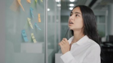 Arabian hispanic businesswoman team leader project manager stand near board write tasks creative ideas corporate goals on paper sticky notes check to-do list organize information managing working data