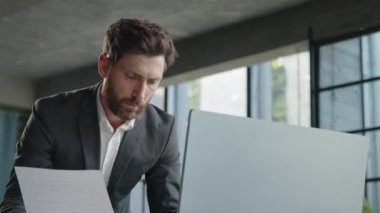 Serious 40s bearded businessman entrepreneur employee standing in office looking at papers report information papers check data in online net service in computer typing working paperwork deadline
