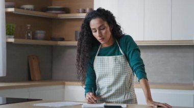 Arabian hispanic housewife pretty curly woman wears apron stand in kitchen write notes food recipe use calculator for counting calories checklist of products calculate domestic bills paperwork at home