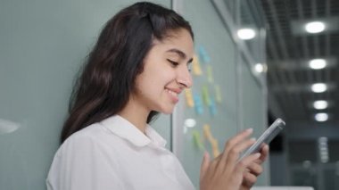 Side view young smiling woman browsing social network makes order in online store using smartphone remote communication in friendly chat on phone female office manager spends free time on telephone