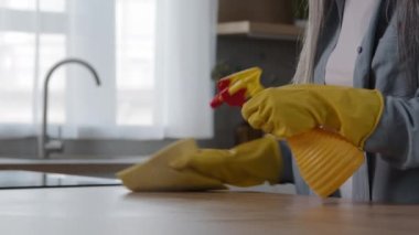 Unknown elderly housewife cleanup worker cleaner granny maid in yellow gloves wash kitchen wooden surface wipe dinner table by wet rag at home keep neat hygiene spraying detergent liquid on countertop
