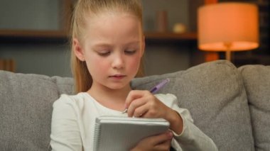 Smart concentrated schoolgirl caucasian child primary school pupil focused writing essay doing math homework sit on sofa making notes in notebook train calligraphic handwriting studying alone at home