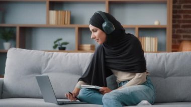Islamic Arabian student girl Muslim woman wearing black hijab busy lady in headphones with laptop watching webinar lesson online class training write notes in notebook do homework e-learning at home