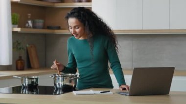 Young housewife woman watch online cookery tutorial on laptop preparing tasty meal checking internet food recipe. Smiling girl mother cooking soup for children use cookbook menu app in modern kitchen