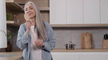 Energetic elderly singer grey-haired old carefree woman lady singing favorite song in wooden spoon domestic karaoke party at home dancing dynamic active moves to music listen loud radio feel amusement