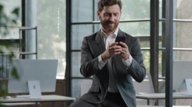 Adult busy bearded 40s businessman boss leader director employee entrepreneur using smartphone worker man browsing mobile phone in office sitting on table working in digital application gadget online