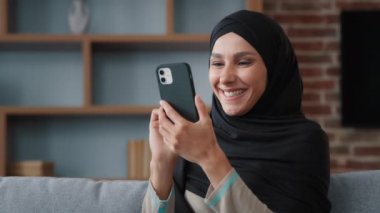 Smiling Arab Muslim Islamic 30s woman user in headscarf black hijab using phone at home sitting on couch middle eastern girl lady holding mobile gadget device smartphone indoors buying online in app