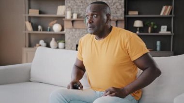 Male bachelor interested shocked man african american older man grandfather sit on sofa in living room watch TV sport program serial series scientific news channel hold hand to chin look at television