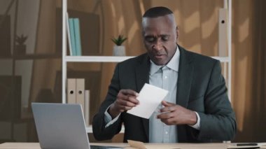 Old senior mature biracial boss company CEO worker amazed shocked African man success businessman sitting at office table open envelope read letter correspondence with good news approval loan credit