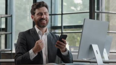 Bearded middle-aged business man in office sitting table using phone mobile audio app service to listening music singing song with modern wireless headphones enjoy online radio website in smartphone