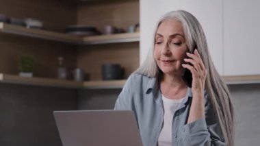 Female senior businesswoman middle aged grey-haired woman specialist freelancer sit in kitchen negotiating with business partner remotely by phone work on laptop e-learning modern tech usage concept