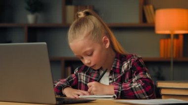 Caucasian kid girl preschool child pupil studying online from home write homework at copybook watch web class lesson learn by video call self-education in pandemic isolation listen educational tutor