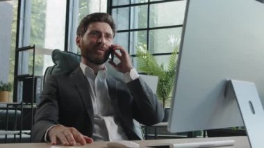 Busy millennial bearded man in office company CEO worker manager consulting client by telephone sitting at table with computer. Professional broker salesman remote mobile phone conversation ecommerce