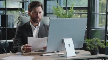 Serious adult 40s bearded pensive thoughtful businessman working at computer in office with paperwork. Professional employee worker typing thinking over online project check data in documents contract