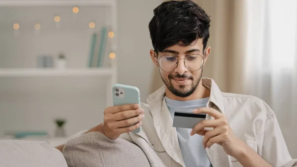 Close-up bearded arabic business man in glasses with credit card and phone makes purchase online shopping enters bank number online transaction uses mobile financial application booking order buying