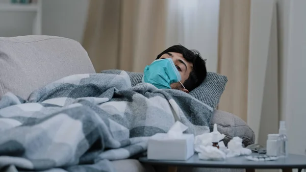 病気に苦しんでいる病気の男体調不良インフルエンザコロナウイルスの症状は 医療用マスクを身に着けています自宅のソファで毛布で覆われている薬波頭の下に隠れて再生されません — ストック写真