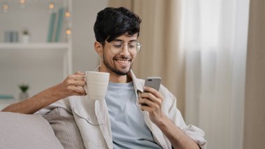 Milenyum Arap erkeği Hintli yakışıklı 30 'lu yaşlarda kanepede oturmuş sıcak çay içiyor fincandan kahve kapuçino kakao içiyor telefonuna bakıyor akıllı telefonlu arkadaşlarıyla sohbet ederken gülümsüyor.