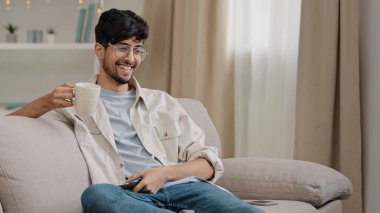 Arab man bearded guy with glasses resting at home sitting on couch drinking tea coffee watching funny movie championship sports game TV rejoices at victory of favorite team laughing fun sincerely