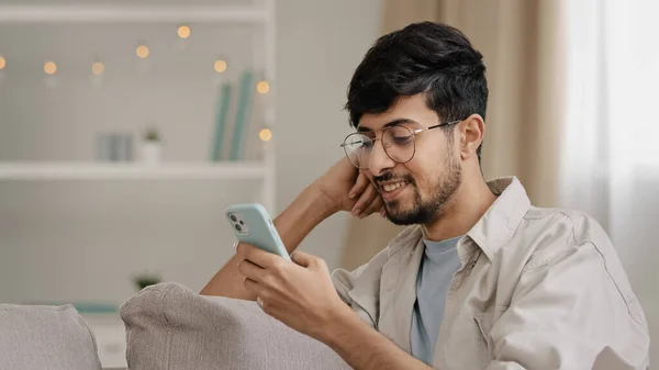 Close-up arabic hispanic guy young man wearing glasses bearded male user blogger sitting at home with mobile phone chatting online browsing web typing message smiling uses smartphone free wi-fi indoor