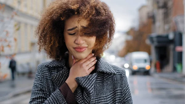Female portrait young sad curly african american girl woman feeling sore throat discomfort coughing suffering from flu symptoms of coronavirus asthma respiratory illness standing in city cold autumn
