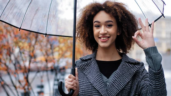 美しい幸せな成功した女性巻きアフリカ系アメリカ人女性の女の子とともに透明な傘スタンドで秋の都市で雨の中でカメラを見て笑顔ショーOkジェスチャー承認サイン同意記号同意 — ストック写真