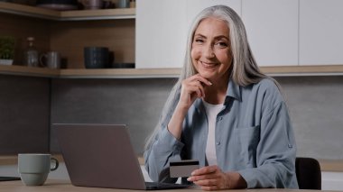 Elderly lady buyer grandmother shopper mature woman sit in kitchen pay utility bills on laptop use banking app for online charity enter security card number at computer internet financial transaction