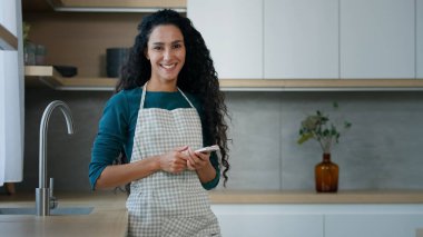 Glad smiling housewife arabian hispanic woman mother homeowner with long curly hair wears apron stand in kitchen chatting sms make cooking notes use mobile phone search internet recipe food delivery