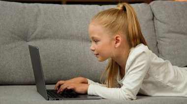 Carefree little kid pretty blonde girl child daughter using laptop lying on grey sofa surfing internet chatting with classmates playing games studying online at home. Children tech addiction concept
