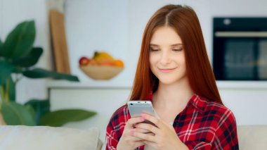 Smiling red-haired caucasian girl model woman sits in kitchen room holds modern gadget smartphone looks at mobile display reads sms messages from boyfriend uses home wi-fi internet plays mobile game