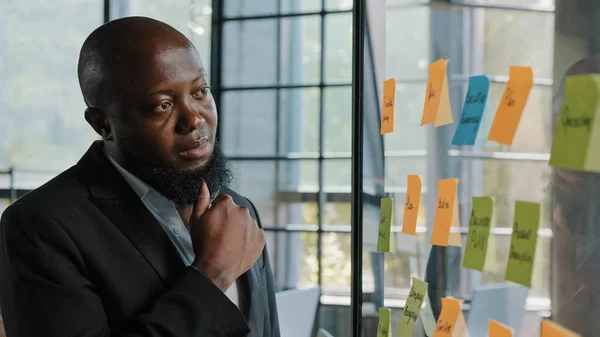 Thoughtful african entrepreneur american adult bearded male businessman team leader stand alone look at transparent glassy wall with paper sticky notes managing goals project planning effective work