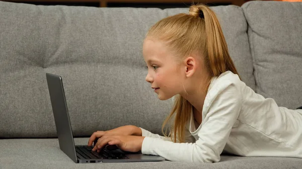 Carefree little kid pretty blonde girl child daughter using laptop lying on grey sofa surfing internet chatting with classmates playing games studying online at home. Children tech addiction concept