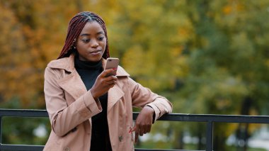Young serious female standing in autumn park holding mobile phone browsing email remotely communicating in social network using smartphone outdoors searching for information on internet writes sms