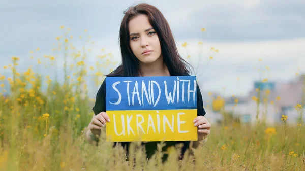 Sad female ukrainian girl migrant woman upset lady posing in field outdoor colorful written cardboard stand with Ukraine slogan demonstrates national patriotism asks help for people solidarity concept