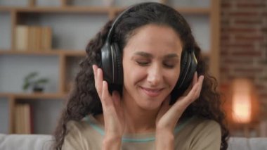 Front view close up smiling Hispanic woman with smiling enjoining lady relaxing at home with closed eyes listening to music in headphones audio sound song feeling well good mood hobby holiday indoors