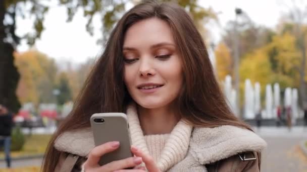 美丽的黑发白人女孩都市女用户站在秋天的城市公园里拿着现代智能手机小玩意用网络图聊天看手机荧幕看有趣的视频在外面消磨时间 — 图库视频影像