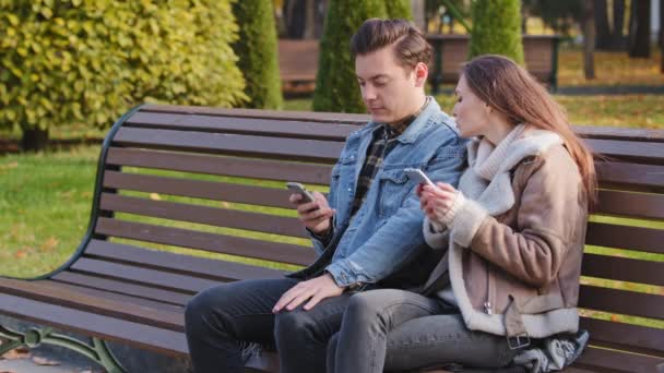 白种人夫妇两个年轻人的伴侣坐在长椅上使用手机小玩艺儿滚动互联网页面查看社交网络显示的图片 彼此在城市公园里度过的时光都有手机成瘾的经历 — 图库视频影像