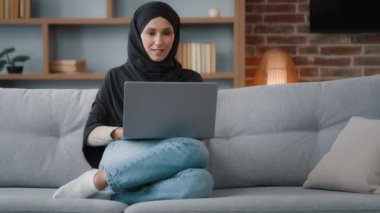 Muslim Islamic woman in hijab sitting at home living room relaxing on sofa couch using laptop online website ordering booking in internet shop working studying remote distant chatting typing computer