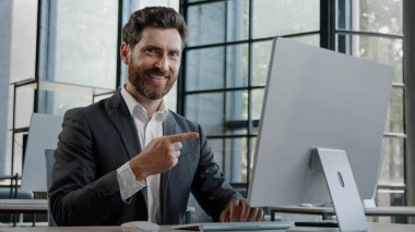 Ofiste çalışan beyaz sakallı bir iş adamı online bilgisayar uygulaması web sitesinde işaret parmağıyla işaret parmağıyla ekranı işaret ediyor.