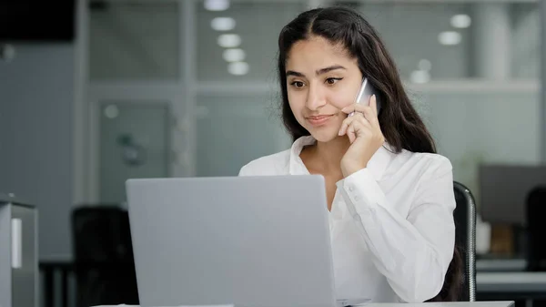 Young arab woman sits in office talks on phone looks at screen laptop makes order in online store orders delivery businesswoman consultant remote advises client on telephone using computer application