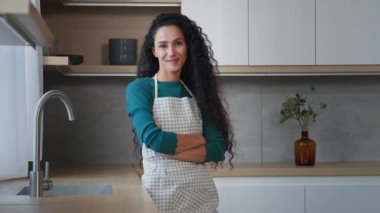 Gülümseyen genç kadın hizmetçi ev hanımı fırıncı kadın uzun kıvırcık saçlı, damalı önlük giyen, modern ev mutfağında poz veren, kendinden emin bir aşçı.