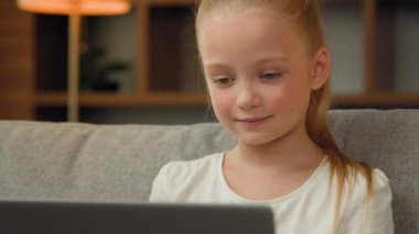 Sevimli küçük kız kanepeye oturmuş modern dizüstü bilgisayar kullanarak çizgi film izliyor komik videolar izliyor bilgisayar ekranlarına bakıp gülüyor ve hafta sonları eğlenceli ev eğlencelerinin keyfini çıkartıyor.